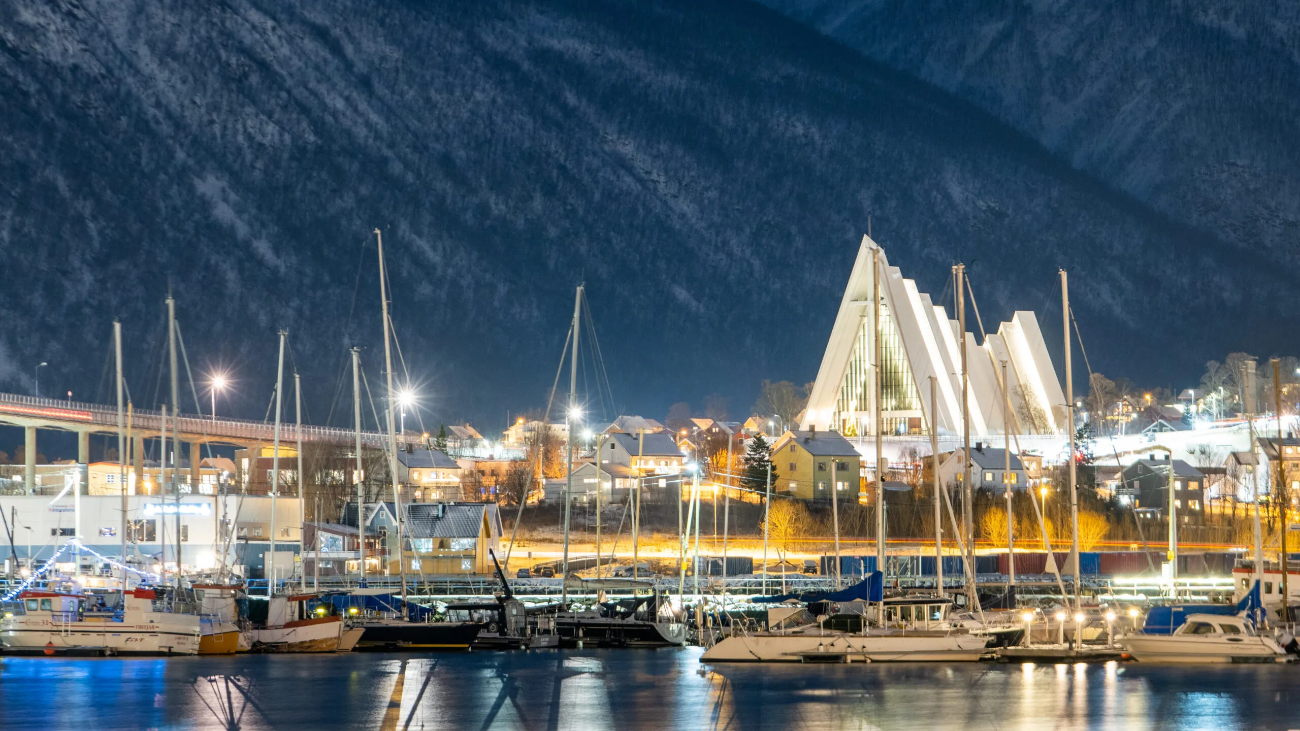 "View of the Arctic Cathedral (Ishavskatedralen) from the harbor in Tromsø, Norway, with its distinctive modernist architecture. The cathedral is set against a stunning backdrop of snow-capped mountains, highlighting the dramatic natural beauty of the area."