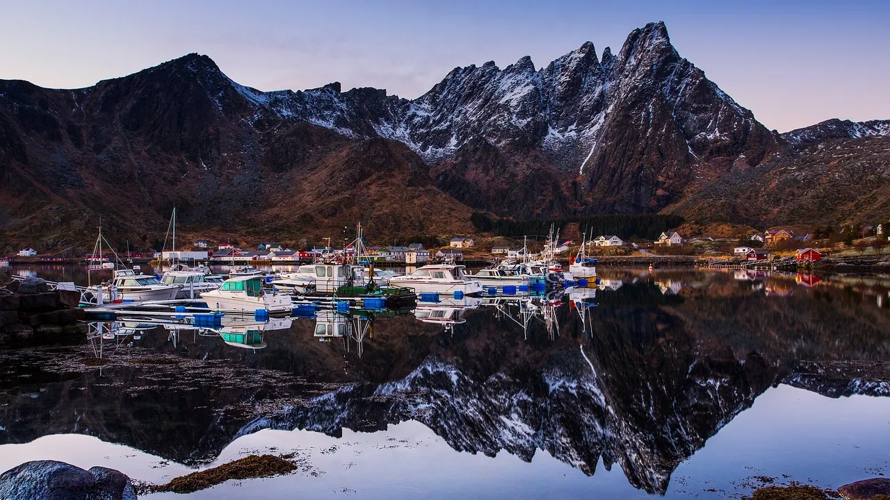 A serene view of a small pier in Ballstad, Norway, jutting out into the calm waters of a fjord, with a majestic mountain rising steeply in the background, reflected perfectly in the mirror-like water.