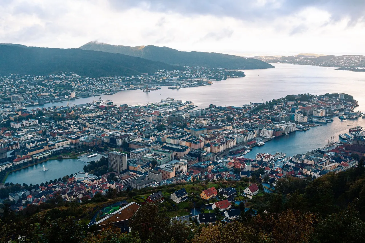 Aerial view of Bergen, Norway, showcasing the city's colorful buildings, historic harbor, and surrounding fjords and mountains, with tiny houses and streets winding through the landscape.