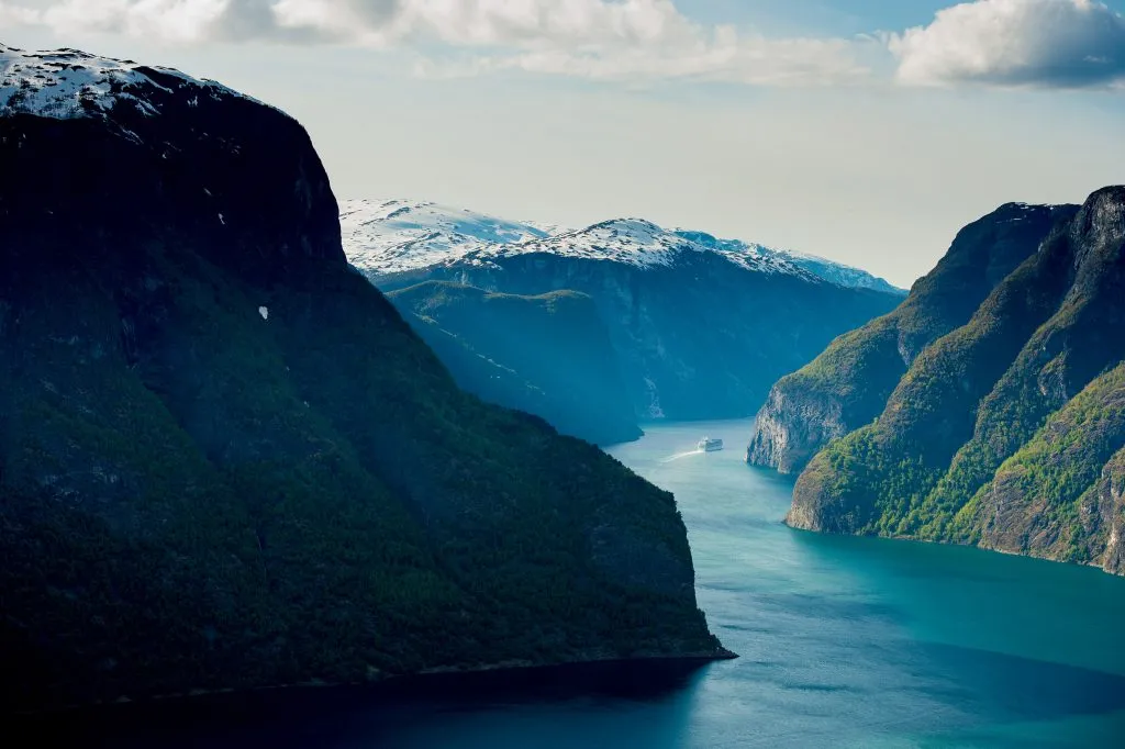 A serene view of a cruise ship navigating through the calm waters of the Aurlandsfjord, a branch of the majestic Sognefjord in Norway, surrounded by steep cliffs and lush green mountains.