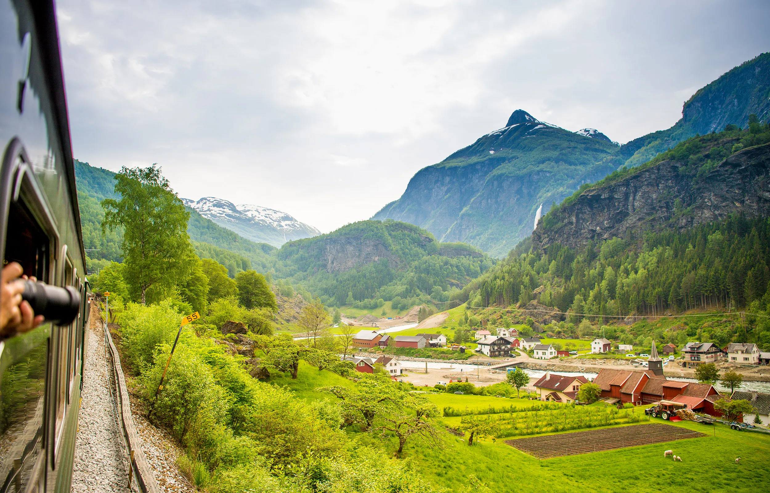 A scenic summer view of the Flåmsbana train tracks alongside the Nærøyfjord, a branch of the Sognefjord in Norway, surrounded by lush green mountains and a few buildings.