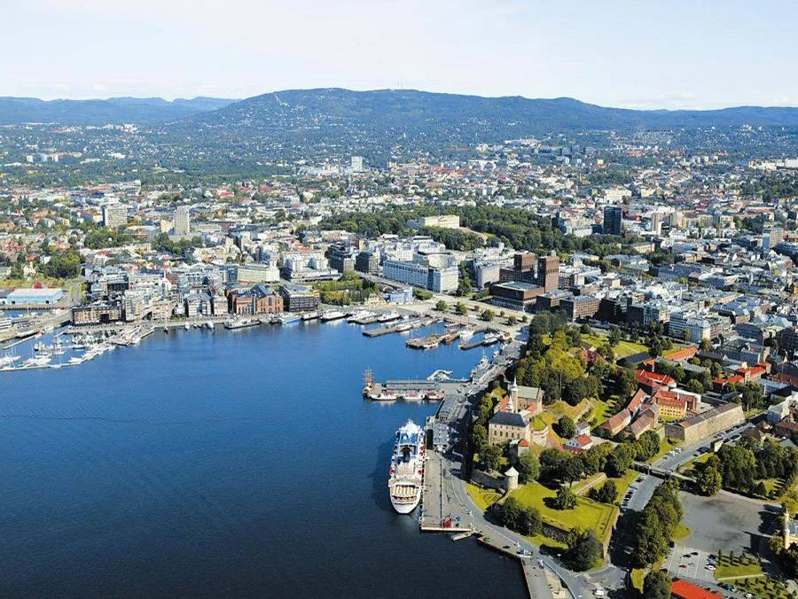 Aerial view of Oslo Harbour, Norway, showcasing the city's vibrant waterfront, with colorful buildings, boats, and ships docked along the pier, set against a backdrop of majestic mountains.